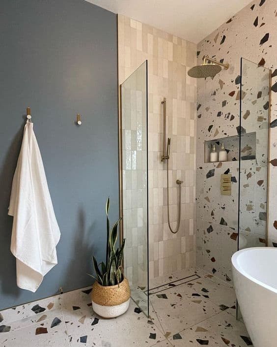 21 STUNNING SHOWER TILE IDEAS FOR SMALL BATHROOMS 21 A chic bathroom with terrazzo flooring, a glass shower enclosure, beige tiles, a freestanding tub, and gold accents complemented by a muted blue wall.