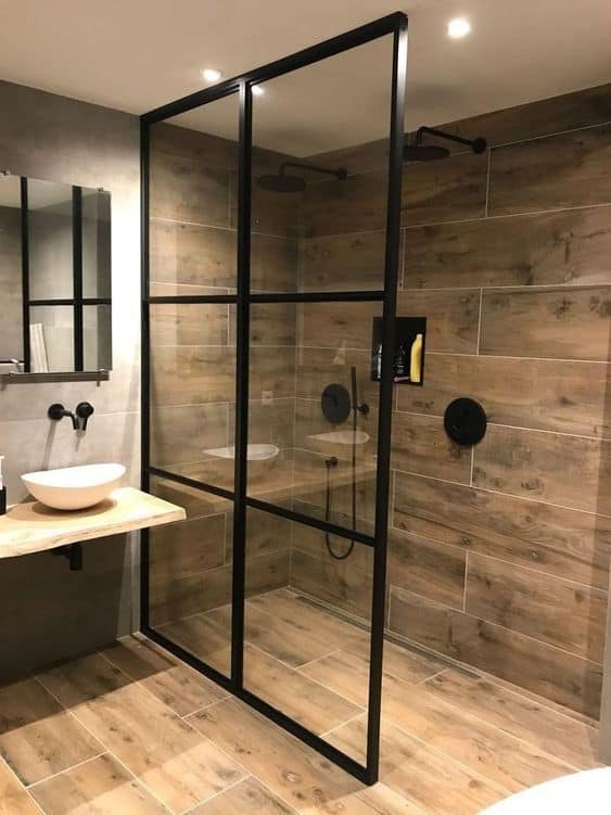 21 STUNNING SHOWER TILE IDEAS FOR SMALL BATHROOMS 9 A modest, rustic bathroom featuring black-framed glass shower enclosure, white vessel sink, and wood-look tiles.