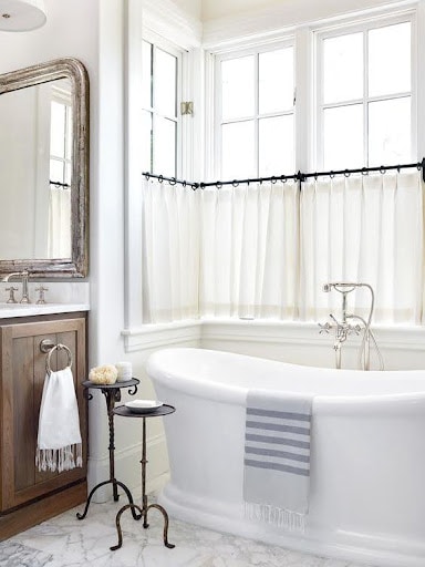 18 SMALL BATHROOM WINDOW IDEAS FOR NATURAL LIGHT AND PRIVACY 3 Vintage-inspired bathroom with a freestanding bathtub, white privacy Cafe Curtains, an antique wood-framed mirror, and bright windows.